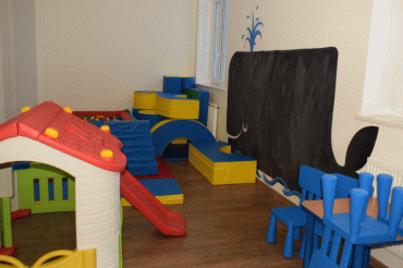 W sali zabaw dla dzieci jest domek, niebieskie krzesełka, a na ścianie namalowany czarny wieloryb.
