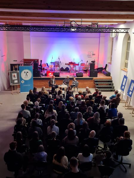 Zdjęcie wykonane z góry. Przedstawia uczestników spotkania "Korzenie", siedzących na krzesłach przed sceną w budynku hali im. Leszka Błażyńskiego w Ełku.